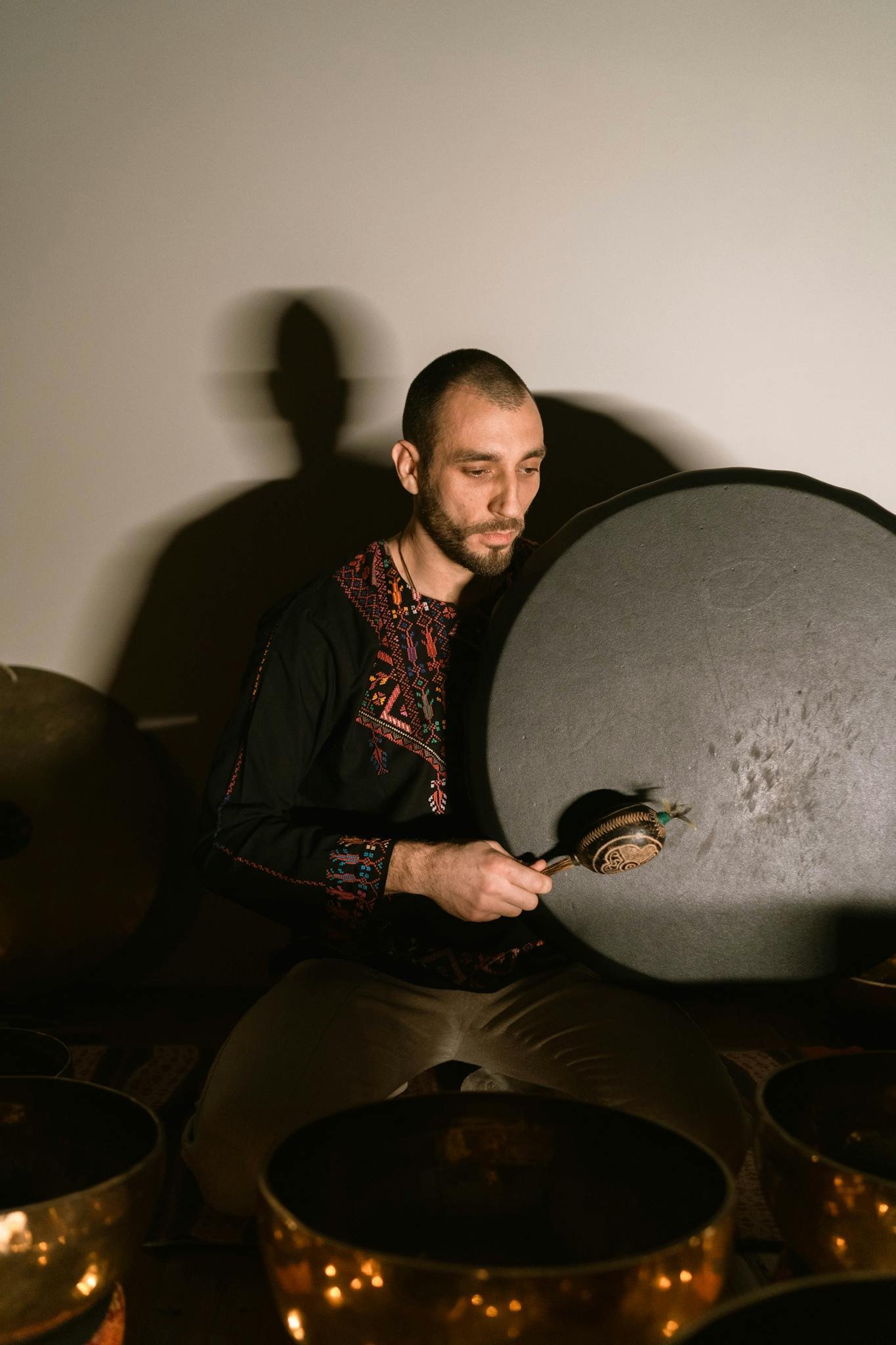 A man meditates indoors with a large gong and Tibetan singing bowls, in a peaceful setting.